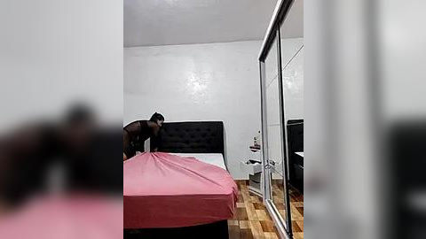 Media: A video of a modern bedroom with a pink bedspread, black headboard, and wooden floor. A blurred figure, possibly a man, stands near the bed.