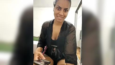 Media: Video of a smiling South Asian woman with medium skin tone, dark hair, and medium-sized breasts, wearing a sheer black robe, sitting on a bed with a smartphone in her hand.