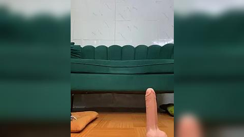 Media: Video of a large, erect penis pointing towards a green, cushioned sofa in a tiled room with a blurred background.