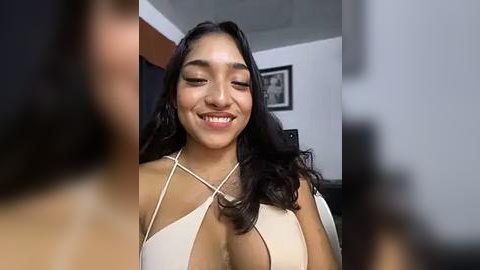 Media: A video of a smiling young woman with long dark hair and medium skin tone, wearing a revealing, beige halter top, taken in a dimly lit room with a framed picture on the wall.