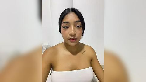 Media: Video of a young Asian woman with medium brown skin, short black hair, and a small tattoo on her right shoulder. She wears a white strapless dress, sitting in a simple room with white walls and a wooden floor.