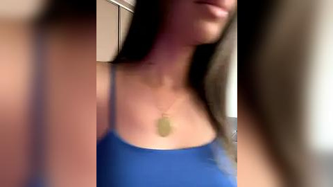 Media: A blurry video of a woman with long, straight dark hair, wearing a blue tank top, standing indoors with a beige wall and a door in the background.