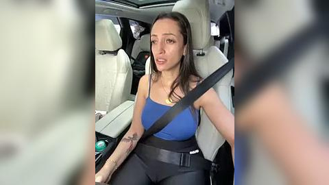 Media: Video of a young woman with medium brown skin and long dark hair, wearing a blue tank top and black pants, driving in a car with beige seats.
