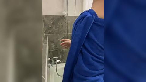 Media: Video of a person, partially visible, in a blue towel, standing in a bathroom with gray marble tiles and a white bathtub.