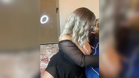 Media: Video of a light-skinned woman with wavy, blonde hair, wearing a sheer black dress, hugging a blue and white pillow, in a dimly-lit room with a circular mirror on the wall.