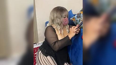 Media: Video of a blonde woman in a black sheer top and striped pants, sitting on a blue couch, holding a phone. Background includes a beige wall and a red carpet.