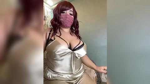 Media: Video of a curvy, light-skinned woman with long, dark brown hair and a pink face mask, wearing a shiny, off-shoulder, gold satin top with black lace, and high-waisted pants, seated on a beige couch in a dimly lit room.