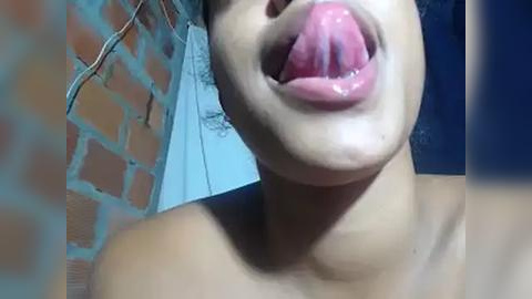 Media: Video of a topless, light-skinned woman with full lips and tongue sticking out, taken indoors against a brick wall and blue curtains.