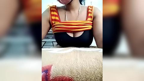 Media: Video of a woman in a black tank top with orange and yellow striped sleeves, revealing her cleavage. She is sitting on a beige carpet, with a blurred staircase in the background.