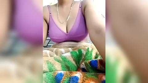 Media: Video of a light-skinned woman wearing a purple bra and a colorful, patterned blanket. She has a necklace on. Background is blurred.