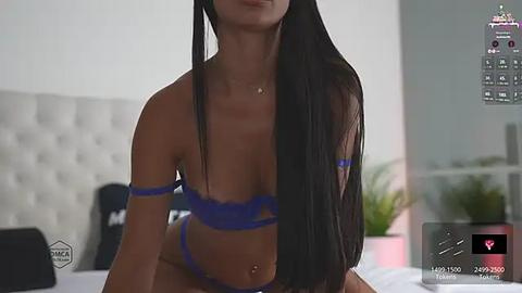 Media: Video of a light-skinned woman with long black hair, wearing blue lingerie, sitting on a bed with a white tufted headboard. The background features a blurred, modern bedroom setting.