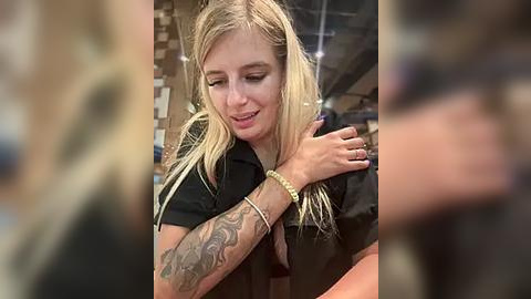 Media: A video of a blonde woman with long hair, wearing a black shirt, embracing another person. She has tattoos on her left arm. The background is blurry, showing a store setting with shelves.