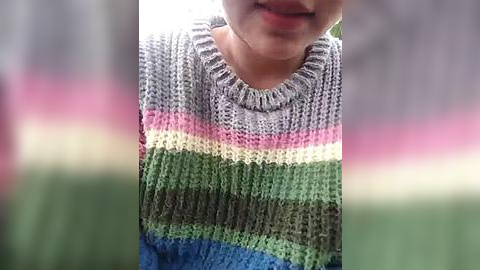 Media: A video of a child's upper torso in a colorful, multicolored knit sweater with horizontal stripes in shades of green, blue, gray, and pink. The background is blurred, featuring a greenish hue, possibly grass. The child's face is partially visible, smiling with closed lips.