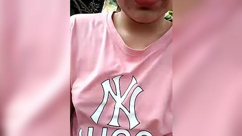 Media: Video of a person wearing a light pink New York Yankees t-shirt with white lettering, partially obscured by pink objects, possibly flowers or a curtain, outdoors.