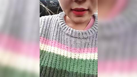 Media: Video of a person with fair skin, wearing a multicolored knitted sweater with pink, green, white, and gray stripes, smiling with bright red lipstick. Background is blurred.