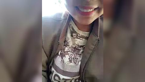 Media: Video of a smiling person with light skin and short hair, wearing a gray jacket over a white shirt with a brown, abstract pattern.