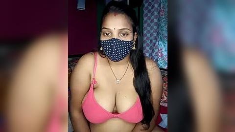 Media: Video of a woman with light brown skin, wearing a pink bra, blue face mask, and a bindi, sitting in a dimly-lit room with colorful curtains.