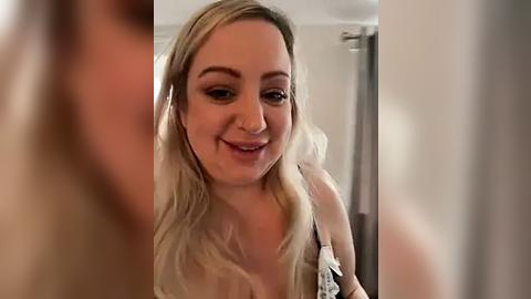 Media: A video of a smiling, fair-skinned woman with blonde hair, wearing a white blouse, standing in a modern bathroom with beige walls and a shower curtain.