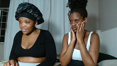 Media: Video of two Black women with dark skin, one with a black satin turban and black long-sleeve top, the other with a high bun and white tank top, sitting on a bed in a dimly lit room.