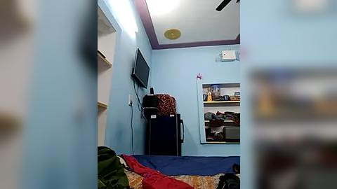 Media: Video of a small, cluttered bedroom with light blue walls and a blue bed, featuring a flat-screen TV, a fridge, a fan, and various items scattered on shelves.