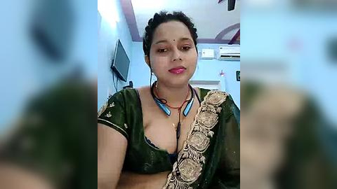 Media: Video of a dark-skinned woman with short, dark hair, wearing a green blouse with a beige floral sari, indoors, with a blurred background.