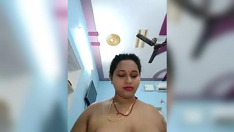 Media: Video of a dark-skinned woman with black hair in a traditional bindi, wearing a gold necklace, standing in a modern room with light blue walls, ceiling fan, and air conditioning unit.