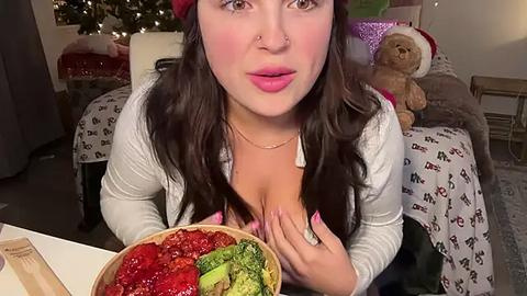 Media: Video of a fair-skinned woman with long dark hair, wearing a white cardigan, holding a bowl of red and green salad. Background includes a Christmas tree, teddy bear, and festive decor.