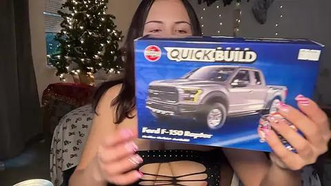 Media: Video of a woman holding a Ford F-150 Raptor toy truck box, smiling, in a cozy room with a decorated Christmas tree and string lights.