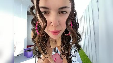 Media: Video of a young woman with curly brown hair, wearing pink earrings, and a nose ring, smiling indoors, with white walls and a purple plant in the background.