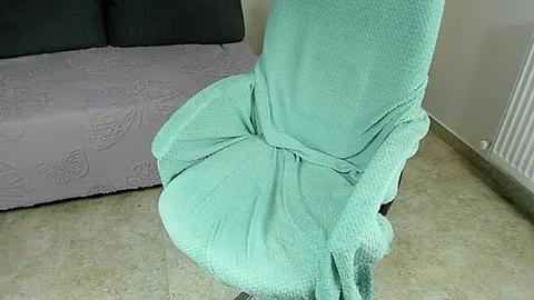 Media: Video of a turquoise, textured blanket draped over a gray couch in a living room with light beige carpeting and a white radiator on the right wall.