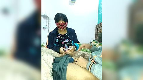 Media: Video of an Asian woman in a red face mask, blue jacket, and floral top, sitting on a bed, holding a phone, with a blurred background of a room.