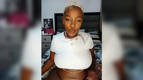 Media: Video of a young Black woman with braided hair, wearing a white crop top, sitting on a bed in a modern bedroom with patterned bedding and gray walls.