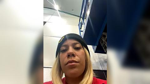 Media: Video of a young, light-skinned woman with blonde hair wearing a black hat, red shirt, and serious expression, taken from a low angle. The background includes blue and white gym equipment.