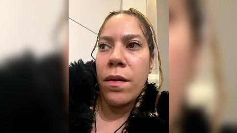 Media: A close-up video of a woman with light skin and braided hair, wearing a black fur-trimmed coat, looking pensive in a neutral-toned room.