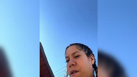 Media: Video of a young woman with braided hair, looking up at a clear blue sky. She has a neutral expression. Background includes a wooden structure with a slanted roof and wire fencing.