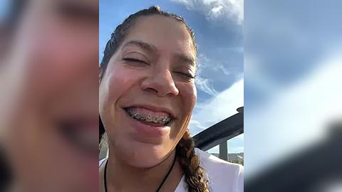 Media: Video of a young woman with a broad smile, wearing a white shirt and a braided ponytail, captured from a car.