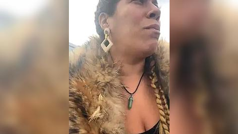 Media: A close-up video of a Native American woman with light brown skin, wearing a fur-lined jacket, braided hair, and a green pendant necklace. She has a serene expression.