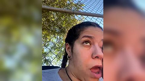 Media: Video of a young woman with dark hair in braids, wearing a gray shirt, captured from the side, looking surprised. Background includes a chain-link fence and green foliage.