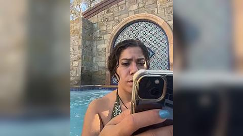 Media: Video of a woman with wet, dark hair, wearing a green bikini, taking a selfie by a pool, with a stone archway and greenery in the background.