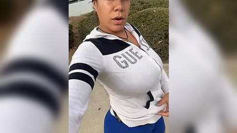 Media: Video of a fit, light-skinned woman with medium-sized breasts, wearing a white \"GUESS\" hoodie and blue pants, standing outside, blurred background, slightly out of focus.