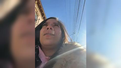 Media: Video of a woman with light brown skin, long brown hair, and a fur-lined jacket, looking up at a clear blue sky with utility lines in the background.