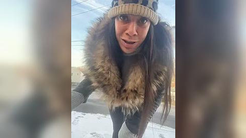 Media: A video shows a young woman with long dark hair, wearing a leopard-print hat, thick fur-lined coat, and grey gloves, leaning against a snowy outdoor railing, with a blurred background.