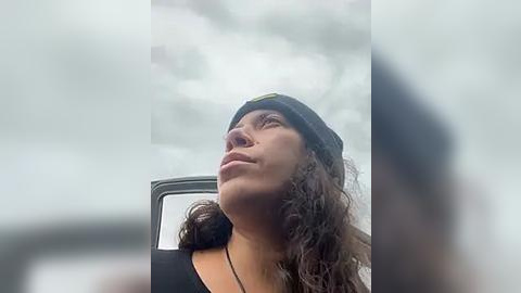 Media: Video of a young woman with curly brown hair and a black beanie, wearing a black top, looking upward with a contemplative expression. Background features a blurred, cloudy sky.