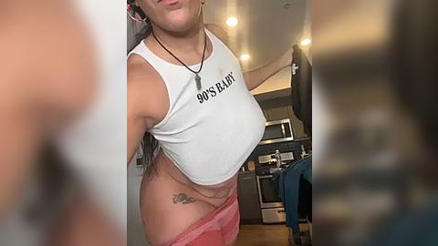 Media: Video of a woman in a white crop top with \"Hooters\" printed on it, pink panties, and a tattoo on her hip, standing in a modern kitchen with stainless steel appliances and a microwave.