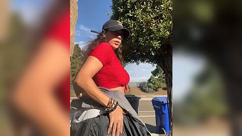 Media: Video of a woman in a red crop top, black cap, and grey skirt, standing by a tree-lined road, with blurred pedestrians in the foreground.