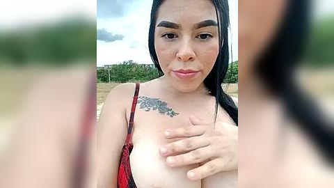 Media: A video of a Latina woman with long black hair and fair skin, wearing a red lace top, holding her large breast. She has a tattoo of a rose on her chest. The background is blurred, showing a grassy field.