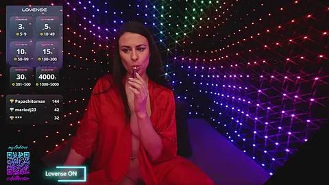 Media: Video of a woman in a red robe, sitting in a dark room with a glowing, colorful LED backdrop. She has a neutral expression and is holding her finger to her lips.