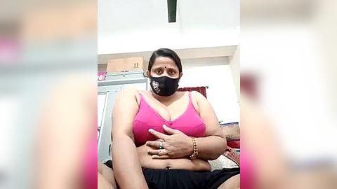 Media: Video of a plus-size woman with medium skin tone, wearing a black mask and pink bra, sitting indoors, holding her stomach.