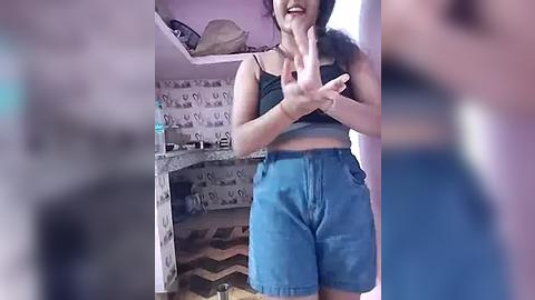 Media: A video of a young woman with a medium build, wearing a black crop top and high-waisted denim shorts, smiling and making peace signs. Background features a bedroom with patterned wallpaper, a bed, and a nightstand.