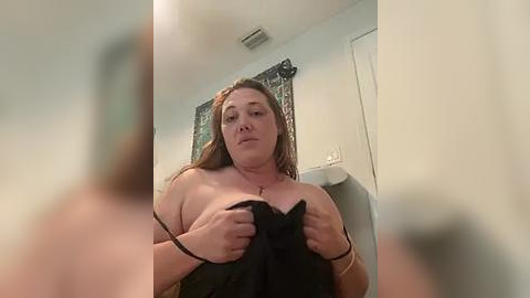 Media: A video of a plus-sized, light-skinned woman with long brown hair, wearing a black bra, standing in a bathroom with white walls, a mirror, and a towel.